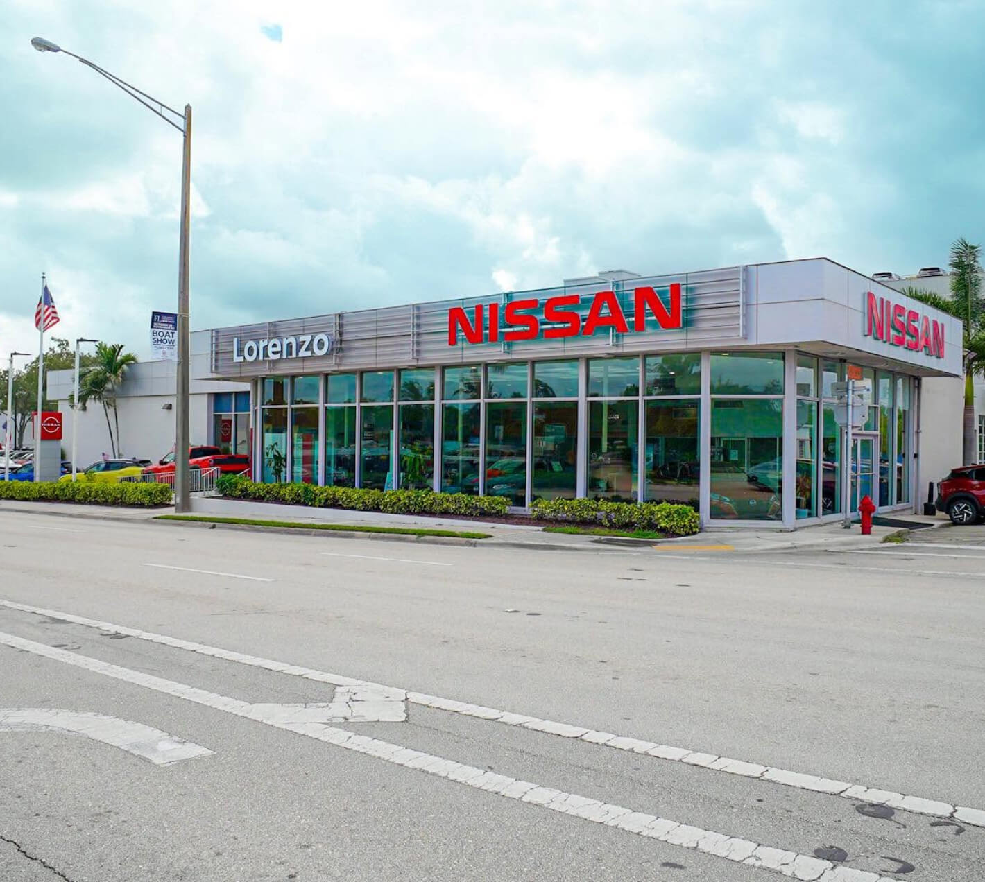 Exterior view of a Nissan dealership facility in featuring a glass-front showroom building with prominent red Nissan signage along the streetfront. 