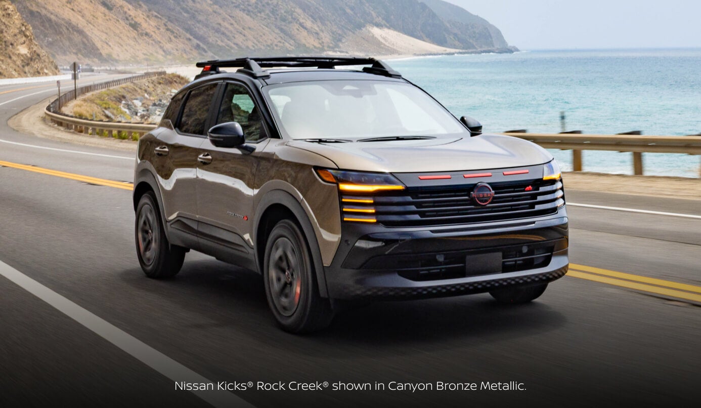 Front three-quarter view of a 2027 Nissan Kicks Rock Creek in Canyon Bronze Metallic driving along a coastal highway beside the ocean.