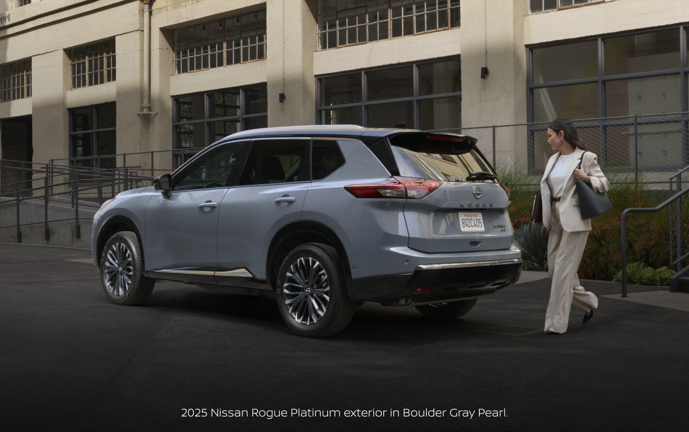 Three-quarter rear view of a 2025 Nissan Rogue Platinum in Boulder Gray Pearl parked beside a pedestrian.