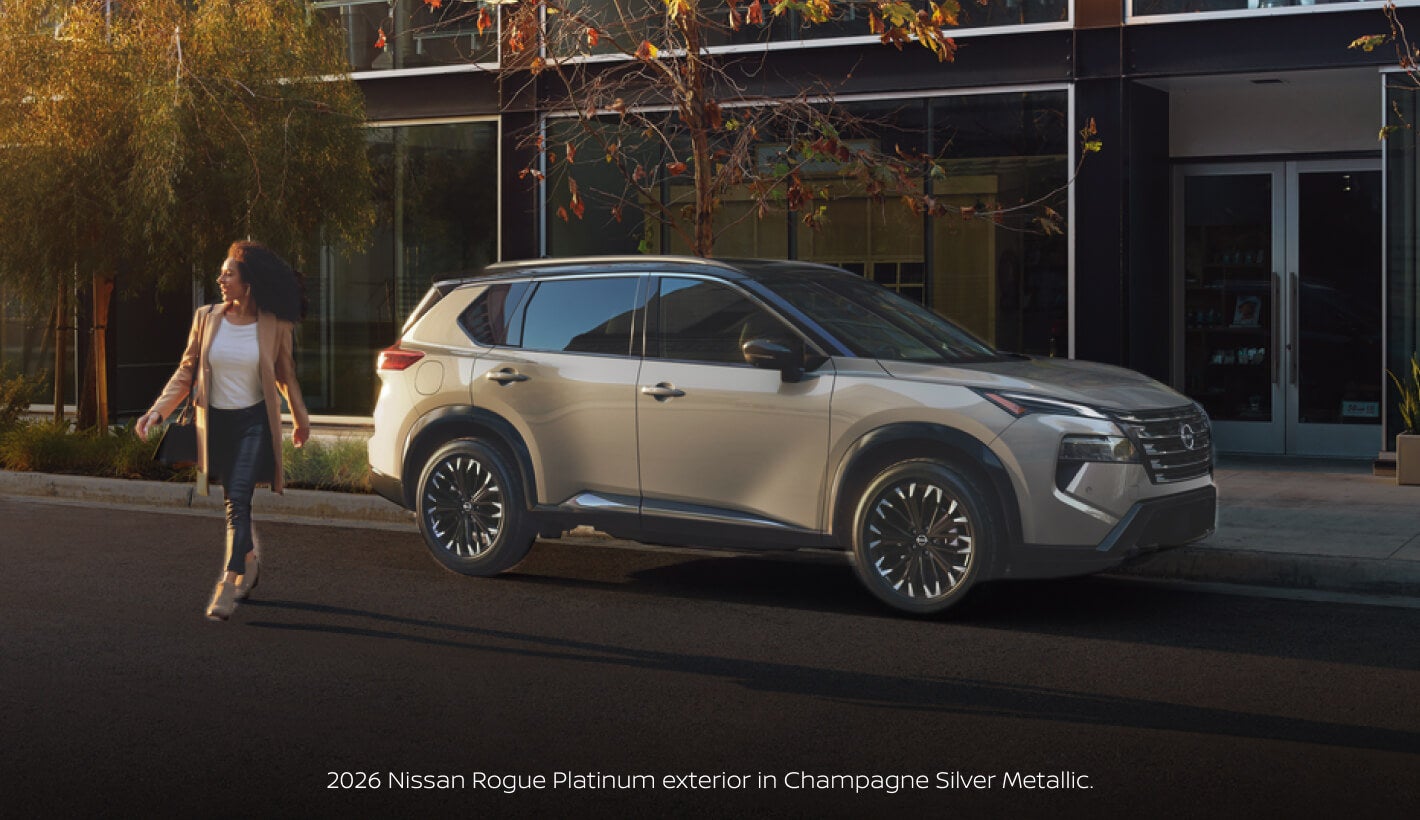 Three-quarter front view of a 2026 Nissan Rogue Platinum in Champagne Silver Metallic parked beside a pedestrian.