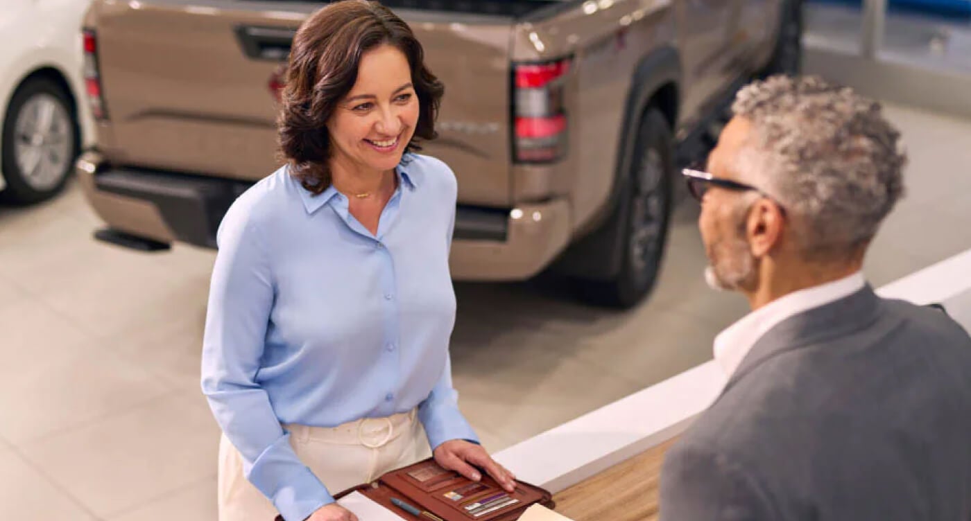A Nissan sales representative in a suit assists a smiling customer in a dealership.
