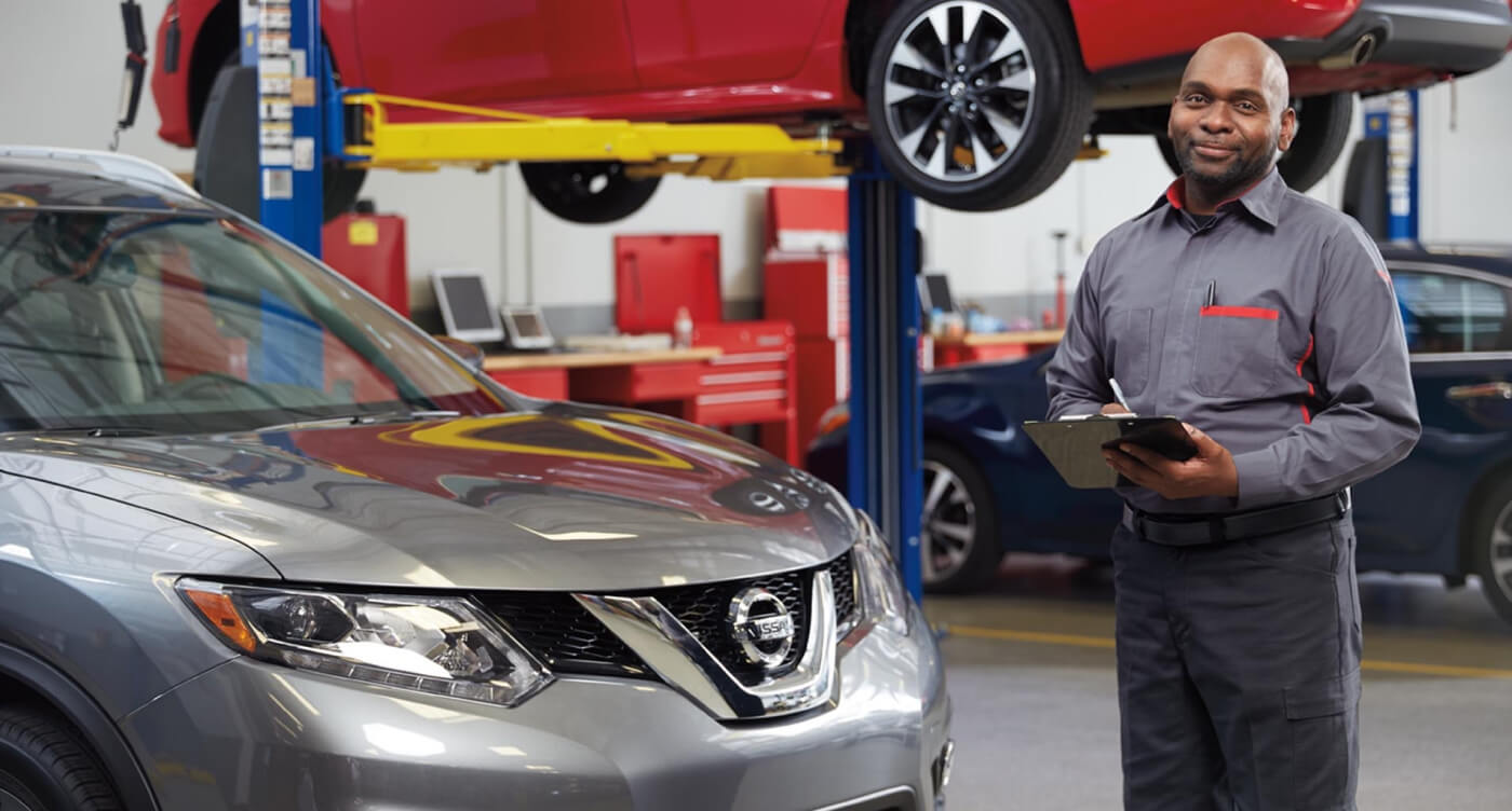 A smiling service technician stands near two Nissan vehicles in a bright service center while holding a clipboard.