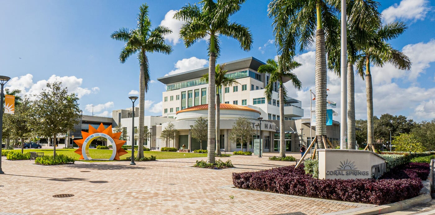 Daytime wide shot of a City Hall surrounded by palm trees, landscaped grounds, and a decorative sun sculpture.
