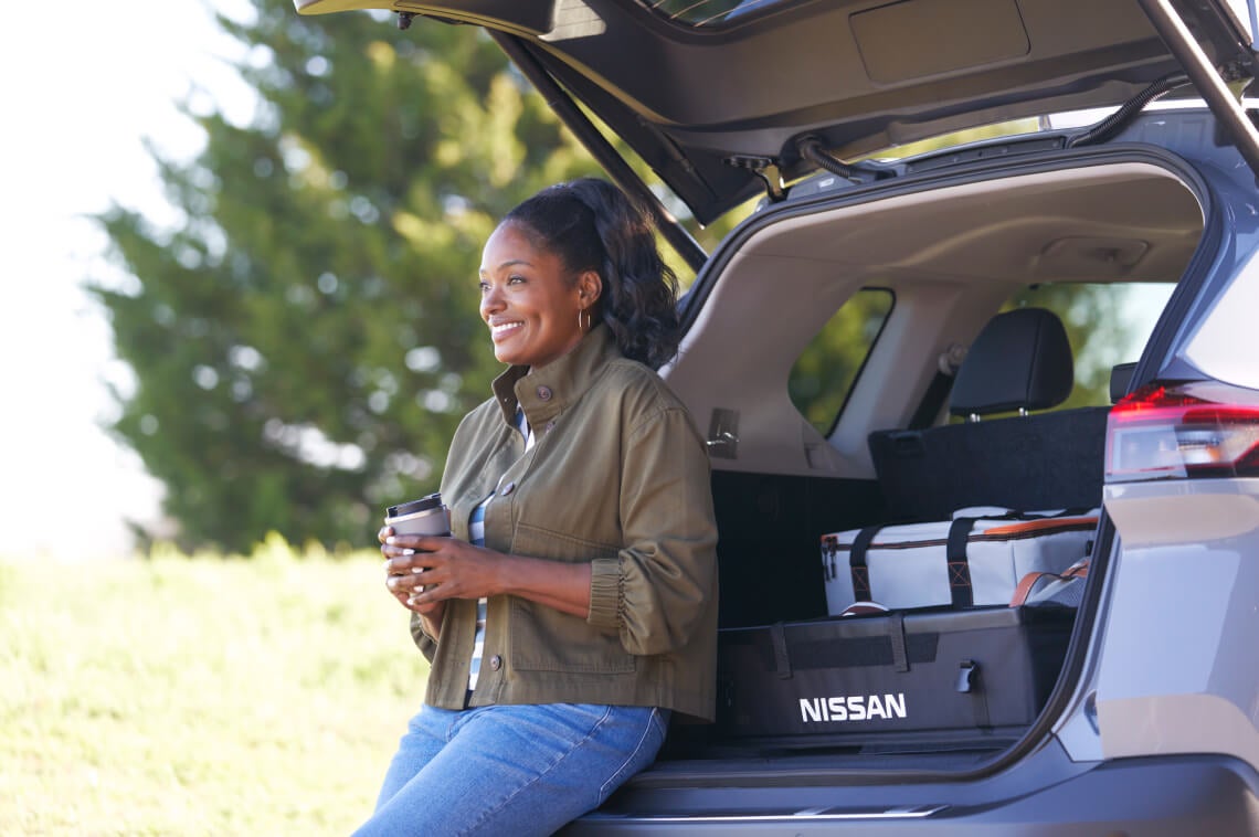 Woman sitting on the edge of her Nissan trunk with coffee in hand
